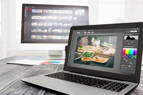 A desktop computer on a desk showing editing software on the screen. In front of it is a laptop showing a photo being edited.