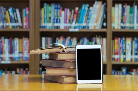 stack of books, tablet on table with bookshelves behind