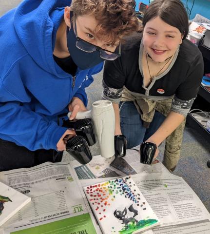 two teens holding multiple hair dryers over a canvas