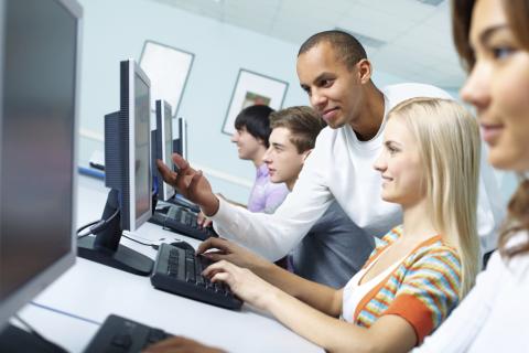 A row of people sit typing at computers. An instructor is pointing at the screen of one of the computers.