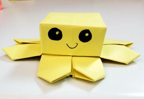 A yellow origami octopus fidget. The head is square shaped and the little critter has big eyes and a smile.