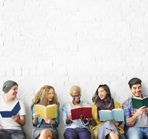 Students, youths, or adults reading seated against a wall. They are talking with each other.