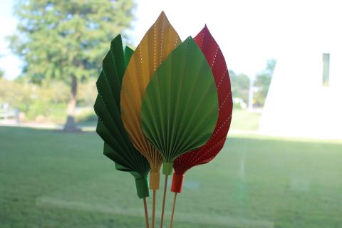 4 paper palm leaves in greens, yellow, and red. Background is a lawn, part of the Joint-Use Library and a tree.