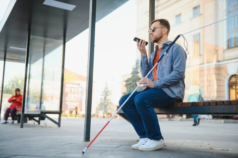 A visually impaired man sits on a bench and speaks to his smartphone.