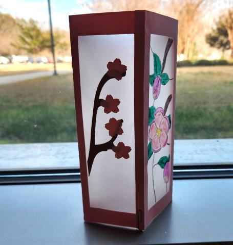 Lantern with paper edges and vellum sides decorated with paper flowers on one side and drawn flowers on another side.