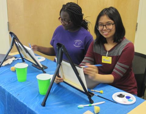 Two teens in front of canvases, ready to paint. One is looking at the canvas, while one is looking up at the camera and smiling.