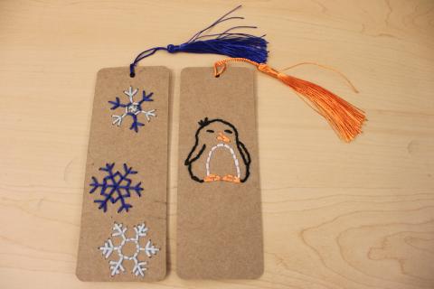 Blue snowflakes embroidered on paper bookmark with a blue tassel. Black, white, and orange penguin embroidered on a paper bookmark with a orange tassel