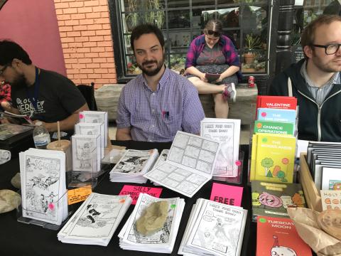 Artist Matt sits behind a table with art spread on it. 