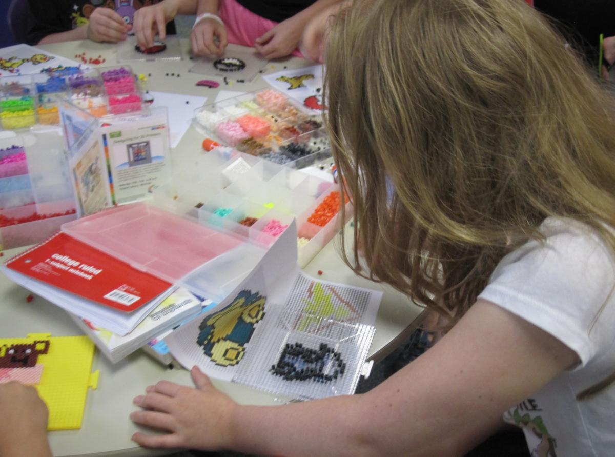 box of Perler beads, teen working on a partially finished design at a table with other teens