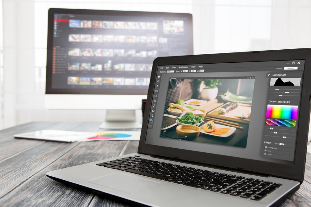 A desktop computer showing a photo library sits on a table behind a laptop that shows photo editing software