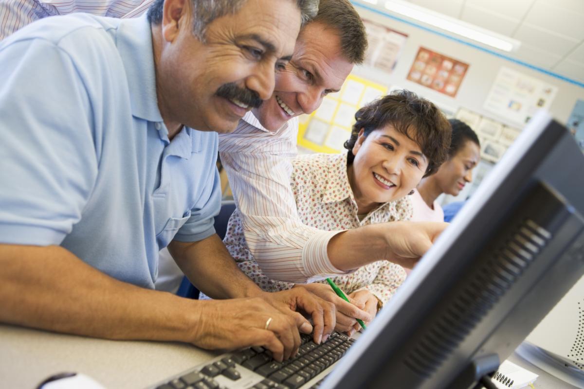 Three adults huddled over a computer, all smiling. A fourth person staring into a computer screen in the background. They are all in a classroom setting. 