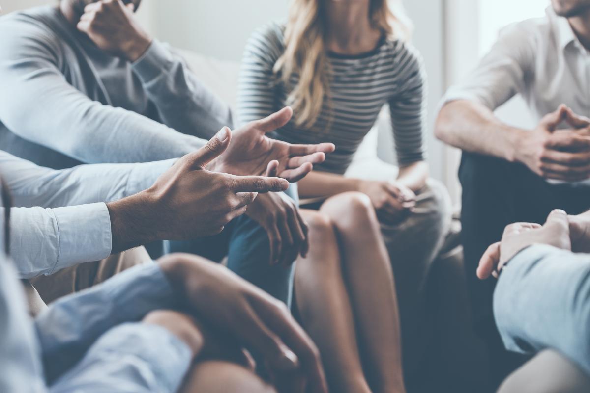 Several people sit in a circle. A pair of hands is being held out in a way indicating that individual is speaking.