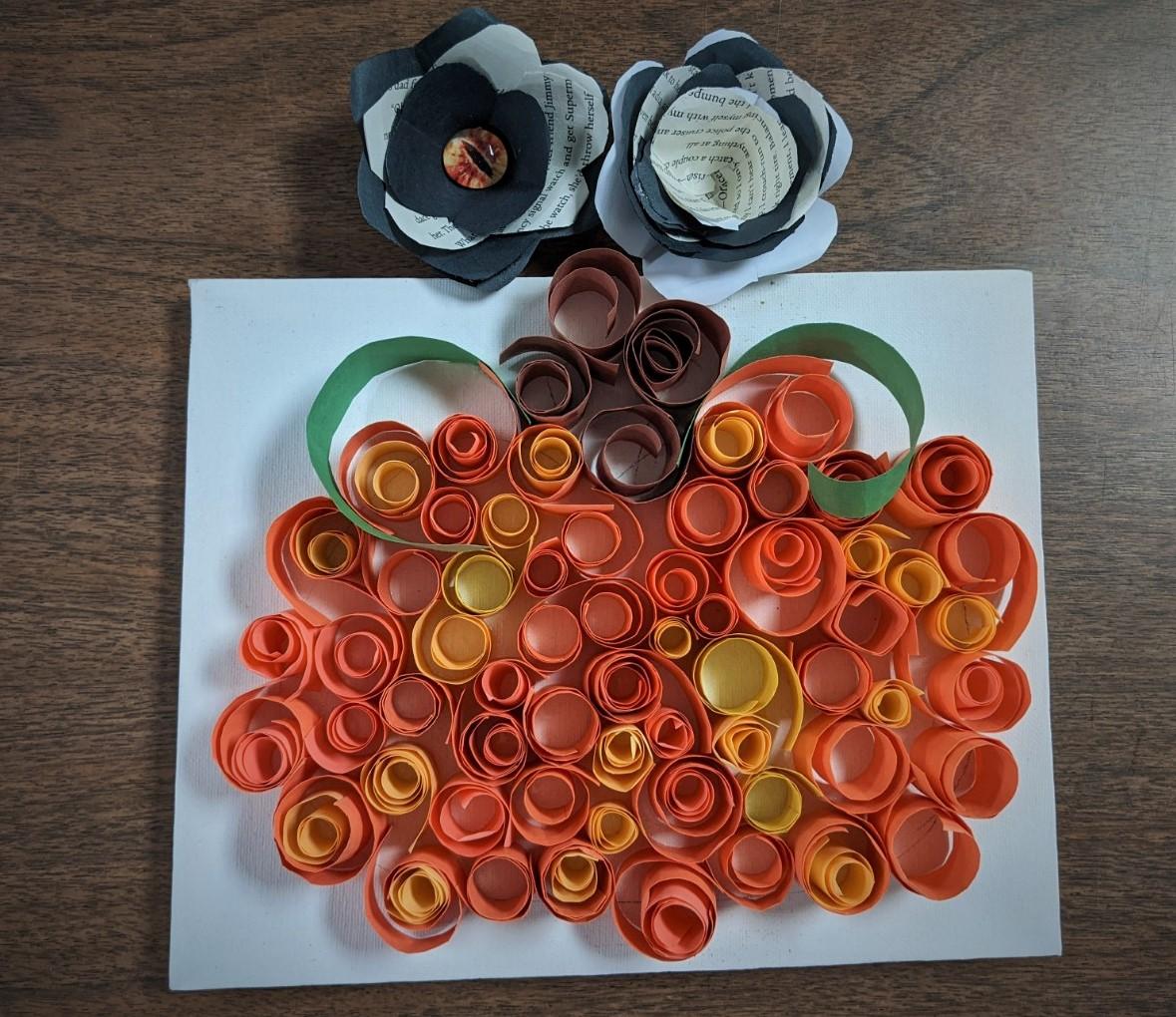 pumpkin on canvas from paper quilling, two book flowers with one having a yellow eye in the center