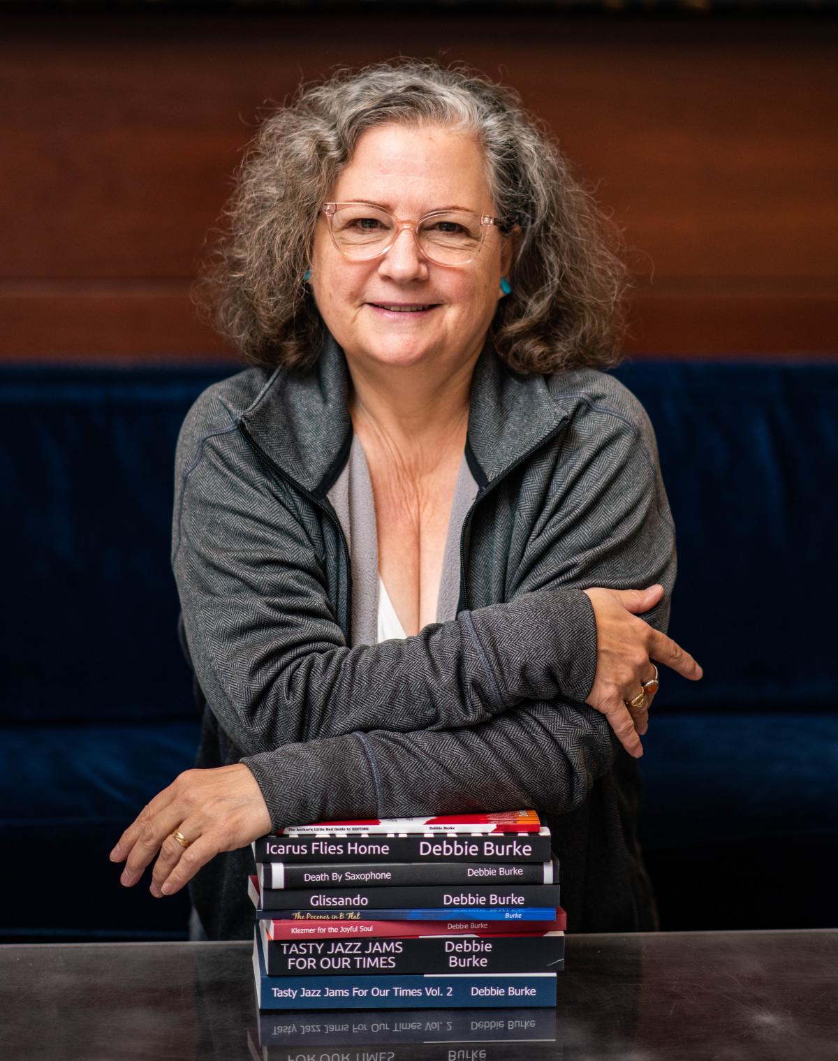 Woman with shoulder length hair and glasses leans on a stack of books