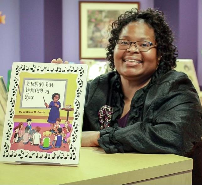 Woman in a black dress sits at a table with a copy of her book on a stand in front of her.