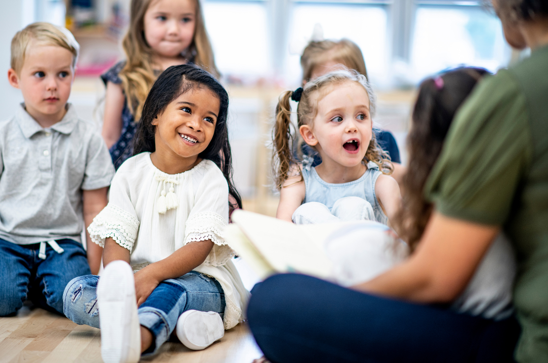 Preschool aged children sitting, gathered facing the teacher who is reading a book for storytime