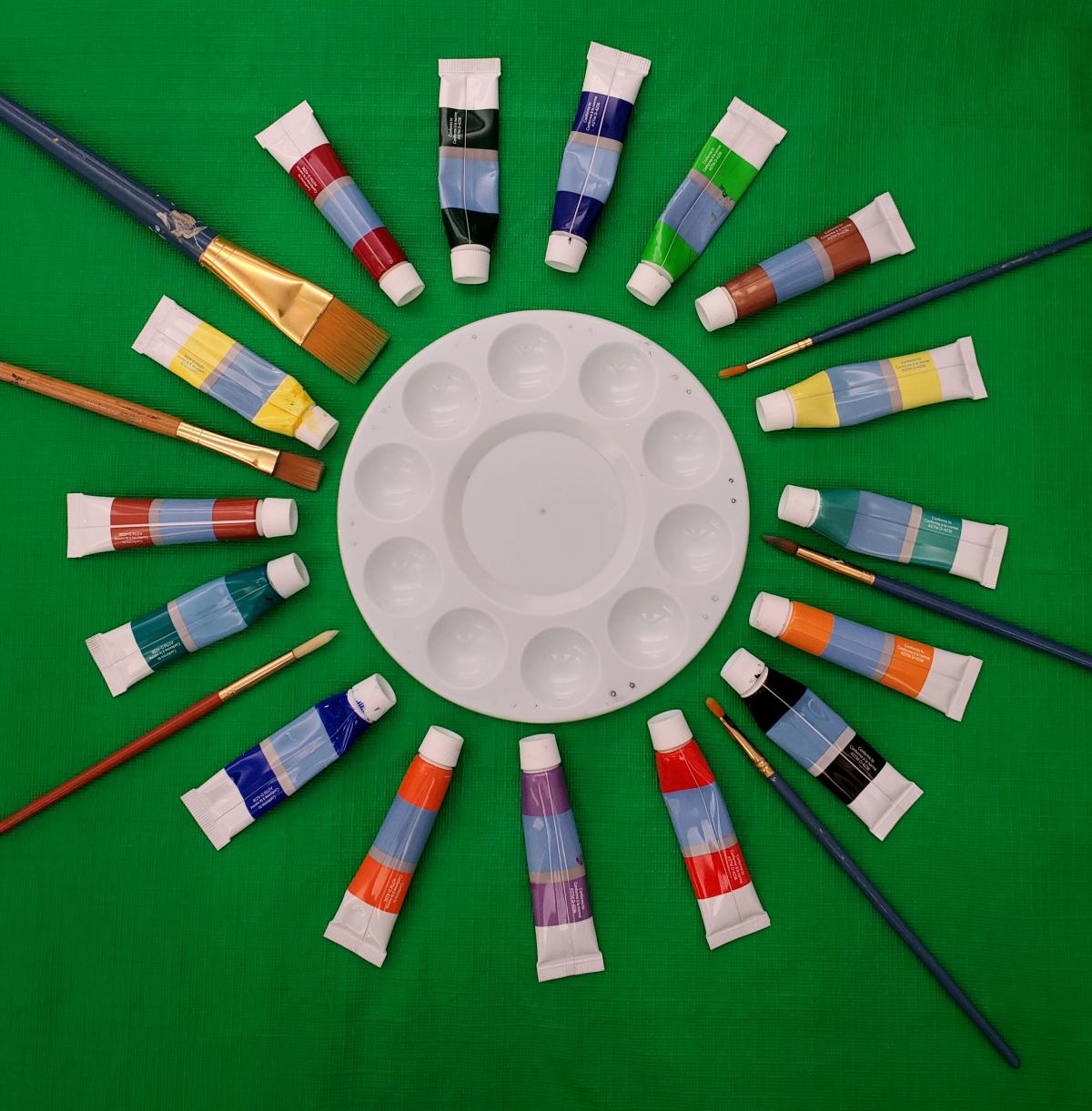 Green background, white paint palette in the center surrounded by colorful paints and paint brushes