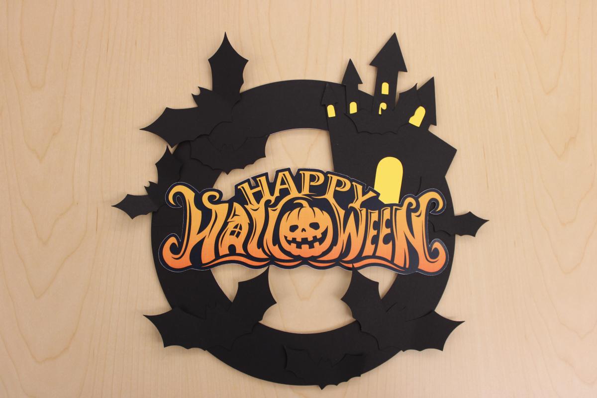 Halloween paper wreath with bats, a haunted house, and Happy Halloween text.